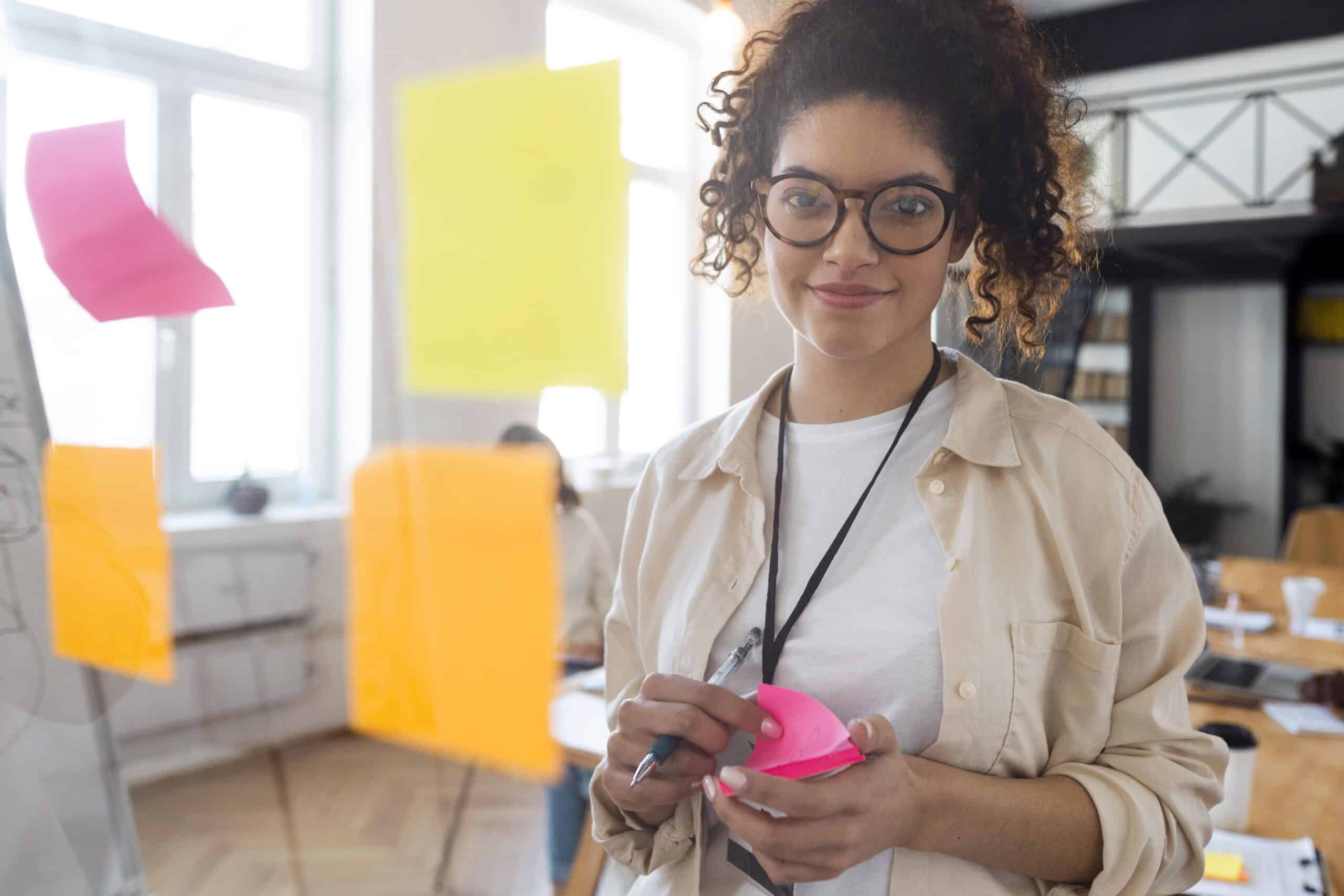 Mulher em ambiente de escritório, segurando post-its e planejando estratégias de marketing digital em um painel com anotações coloridas ao fundo.